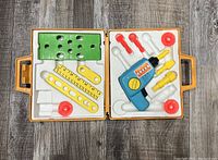 Top view of toy tool kit opened showing blue/yellow drill, yellow rulers, green perforated board, red knobs, and other plastic pieces arranged in molded tray inside yellow carrying case.