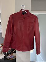 Front view of red leather ladies jacket on hanger, showing zipper front, stand collar, and smooth leather material.