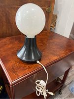 Mid-century modern table lamp with black flared base and large white globe bulb on top, placed on wooden furniture with white power cord