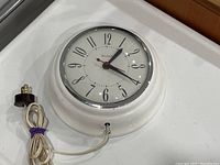 Vintage United electric wall clock with white round metal case, black hands, and attached white power cord with brown two-prong plug.