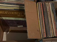 Photo showing box of vinyl records with the album cover "Great Country of the '70s" prominently visible with a rustic wooden barn and wagon wheel graphic.