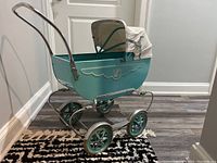 Side view of blue metal vintage doll pram with curved chrome handle and four wheels, fabric shade partially open.
