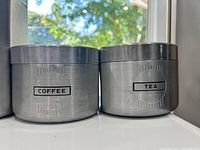 Two smaller metal canisters labeled Coffee and Tea, silver finish, brushed metal, cylindrical shape, black printed labels.