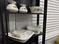 Full view of the 8-piece Corning Ware set displayed on a dark shelf. Includes coffee pots, casserole with lid, rectangular baker and additional bakeware pieces all with floral fruit motif.