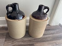 Two vintage stoneware crocks side by side on wooden floor against white wall, both with beige lower bodies and dark brown glazed tops with handles.