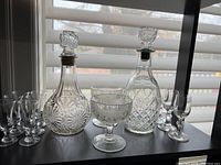Full set of two decanters, six small cordial glasses, four goblets, and two coupe glasses displayed on a dark surface near a window with blinds.