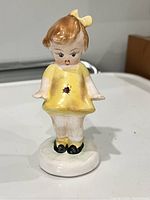 Front view of a vintage ceramic figurine showing the girl’s face, yellow dress with ladybug, black shoes, and white base.