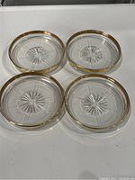 Four clear glass coasters with a starburst design in the center and gold rims around the edges, placed on a white surface.
