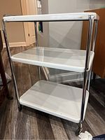 Photo of a vintage medical cart with white enamel shelves and chrome frame against a wooden floor.