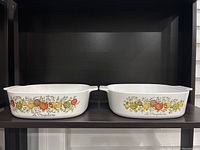 Set of two vintage Pyrex La Marjolaine casserole dishes side by side showing the vegetable and herb designs and handles on each dish.