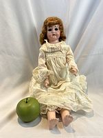 Full view of the antique German doll sitting upright beside a green apple for size reference.