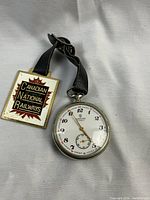 Pocket watch and enamel medallion on leather strap laid flat on white background