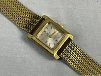 Close-up of watch face and part of mesh bracelet showing silver dial, baton markers, gold tone hands and textured gold-plated case sides.