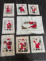 Eight cross-stitch Christmas-themed fabric pieces laid out showing seven different Santa figures and one cardinal bird design