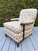 Front angled view of antique carved wooden armchair with floral upholstery on a wood deck with greenery background.