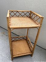 Front angled view of vintage two tier bamboo shelving unit showing woven cane shelves and diamond pattern bamboo railing.