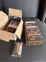 Photo showing two cardboard containers holding multiple cassette tapes, one open box with several cassettes and the other a wooden TDK holder with cassette tapes inside it. One cassette tape is placed outside the boxes on the floor.