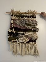 Front view of the handmade wall hanging showing its various wool textures, earthy color palette, and bamboo mounting stick.