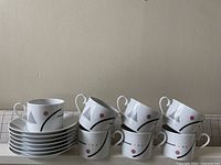 Six white ceramic plates stacked with six matching mugs on top, all showing geometric abstract designs in pink, black, gray, and orange lines.