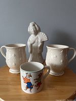 Front view showing Benihana Geisha ceramic tiki mug and three other mugs on table against wall