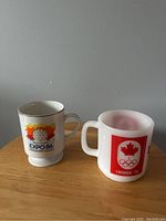 Two mugs on a wooden table, one Expo 86 logo mug and one Canada 76 Olympics mug