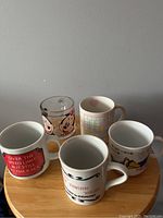 Photo showing 5 assorted mugs on a wooden surface, including two glass Mickey and Minnie mugs and three white ceramic mugs with various prints and slogans.