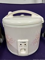 Front view of white Tiger brand rice cooker showing floral print, front panel with cooking switch and handle on lid.