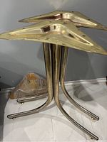Three gold tone metal table legs stacked vertically showing the triangular mounting plates at the top, and tubular curved legs with some tarnishing and fading.