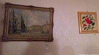 Two framed art pieces hanging on a wall; larger cityscape by Pivos to left and smaller red floral print to right.