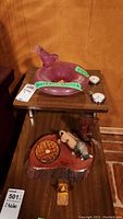 Photo showing purple decorative ashtray with bird figure, two pocket watches on a wooden shelf above a wooden base with ceramic figurine and basket.