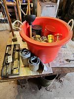 Large red plastic bucket filled with containers and jars, string and other items on top of a wooden bench.