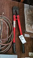Bolt cutter with red handles and black grips shown from above with surrounding items on wooden surface.