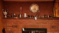 Full view of assorted brass decorative items, ceramic vase, pewter mug, candle, and trophy on brick fireplace mantel.