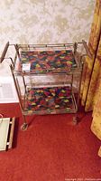 Full view of the two-shelf metal serving cart with stained glass patterned shelves and caster wheels on a red carpet