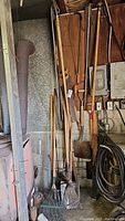 Photo of assorted garden tools hanging from rack and resting on floor in garage, including shovels, rakes, saws, gardening hoe, nails and a black cable coil.