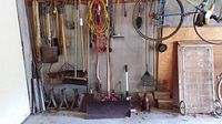 Photo of various garden tools and equipment hanging and stacked against a white wall in a garage, including shovels, rakes, brooms, ropes, car stands, hand saws, a window snow cleaner, wood pieces, and a two wheel cart.