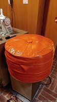 Round orange vinyl mid-century modern ottoman with decorative stitched pattern and central button tuft photographed from side angle.