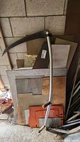 Photo showing assorted flat sheet metals and metal bars leaning against a wall along with a metal scythe with wooden handles in front.