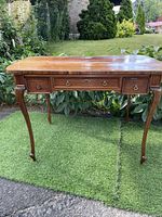 Front view of antique Queen Anne style wooden desk with four drawers and brass handles, placed outdoors on grass.