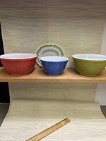 Three Pyrex mixing bowls in red, blue, and green colors with a Spring Blossom gravy boat saucer placed behind them on a wooden shelf with a ruler below for scale.