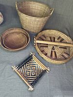 Overview photo showing all five wicker items on a grey fabric backdrop, including two round baskets, one large handled tray, one tall round basket, and one small square wall decor piece.