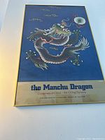 Framed poster featuring a colorful Manchu dragon on deep blue background with exhibit title along bottom. Brushed gold frame and glass visible.