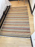 Full view of the rectangular hand woven wool rug on light wood flooring next to bed frame.