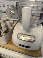 KitchenAid food processor base with clear work bowl attached, control buttons on front, and blade accessories stored in a container to the side.