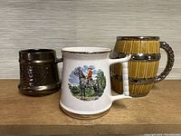 Three vintage mugs placed side by side on a shelf: a brown Prinknash Abbey mug, a white Mountie Prince William mug with horse rider graphic, and a barrel-shaped mug with handle.