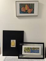 Three framed artworks of fruit: colorful pears in silver frame on wall, black framed large pear print, and rectangular black framed green apples print on table