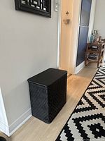 Side view of the vintage black woven laundry hamper against a wall near a patterned rug.