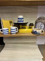 Photo showing TG Green Cornishware flour shaker, yellow Oxfordware water pitcher, Carrigalware mug and creamer, weighing scale with weights, ceramic bread and bun warmer, and a Paul Stories soup mug.