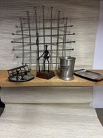Front view of all six metal vintage items displayed on a wooden shelf with neutral background: chrome net fruit bowl, wire biplane sculpture, wire Don Quixote sculpture, pewter julep cup, stainless steel coasters, and ring tray.