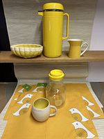 Overall display of all yellow-themed items arranged on a wooden shelf including pottery, carafe, jug, mug, juice jug, and placemats.